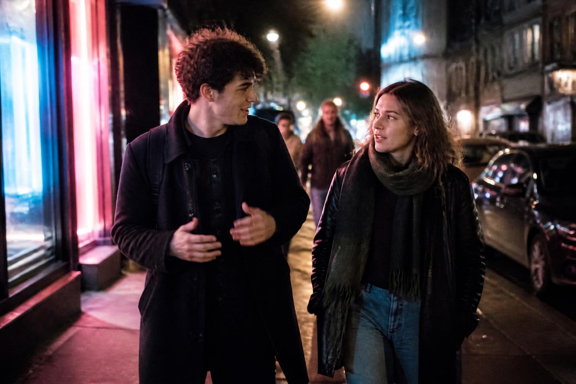 Two students walking and talking on a neon-lit city street at night
